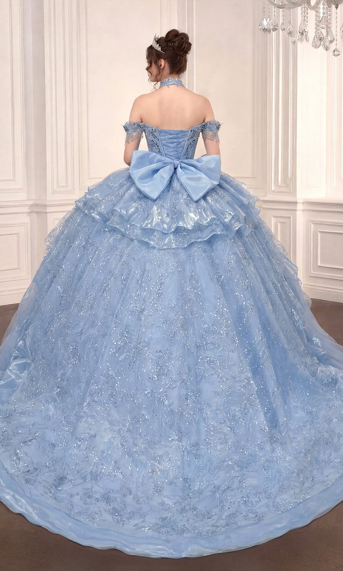 A woman wears the Quinceanera Dress 15742 By Ladivine, a pale blue off-the-shoulder gown with a large back bow, standing in an elegant room with white paneled walls and a chandelier.